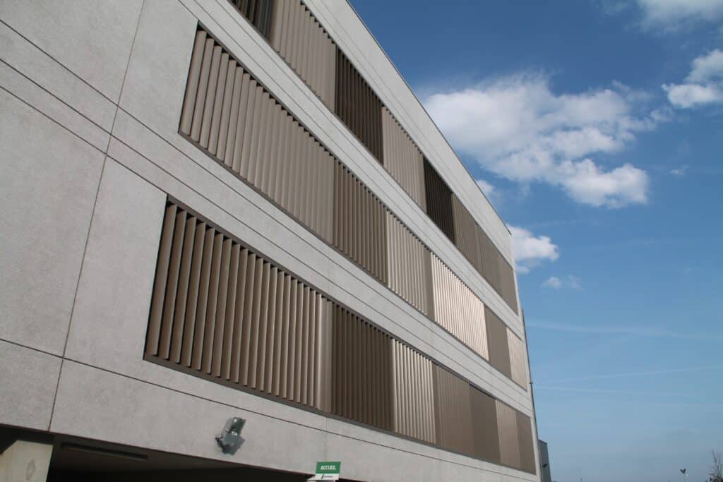Façade moderne d'un bâtiment gris avec des rangées de persiennes verticales marron sous un ciel bleu nuageux. Panneau "ACCUEIL".
