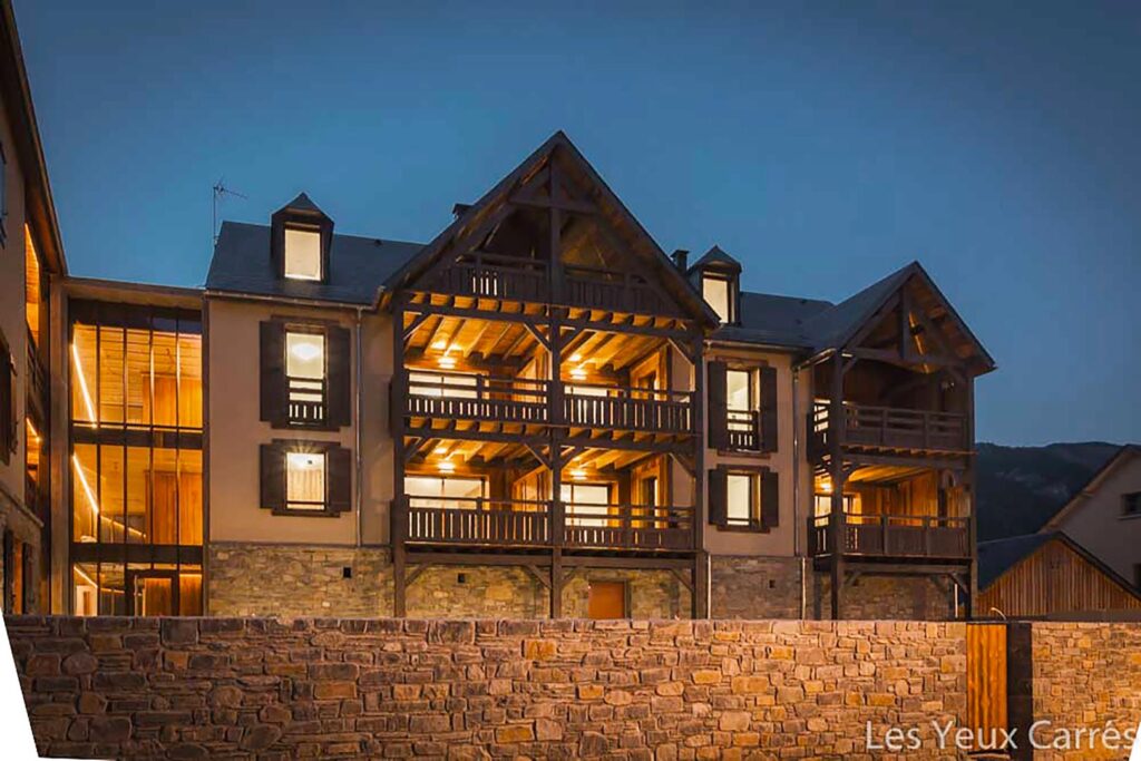 Bâtiment alpin moderne en bois et pierre éclairé au crépuscule. Nombreux balcons et fenêtres lumineuses. Texte : Les Yeux Carrés.