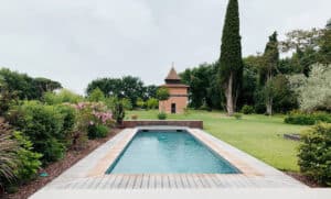Piscine rectangulaire avec deck en bois et carrelage clair dans un grand jardin verdoyant. Une tour en briques au toit conique se dresse au loin.