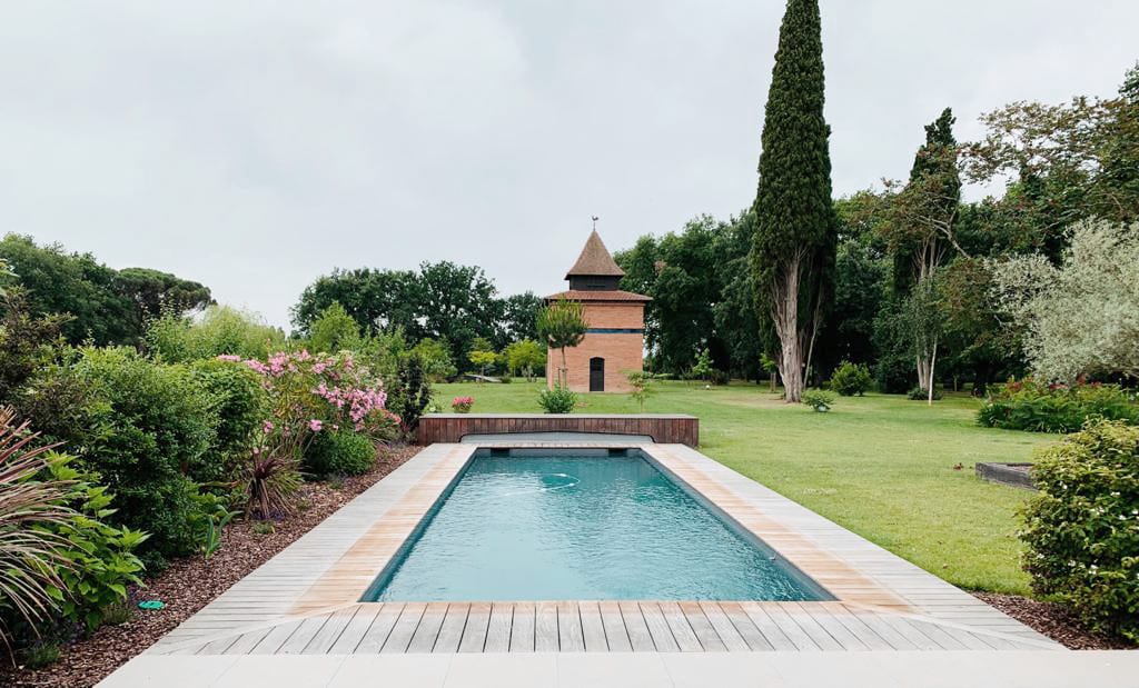 Piscine rectangulaire avec deck en bois et carrelage clair dans un grand jardin verdoyant. Une tour en briques au toit conique se dresse au loin.