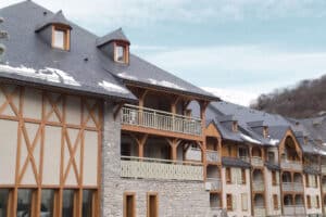 Résidence style chalet en montagne, façades pierre et bois, toits d'ardoise enneigés. Balcons et lucarnes ornés, ambiance hivernale.