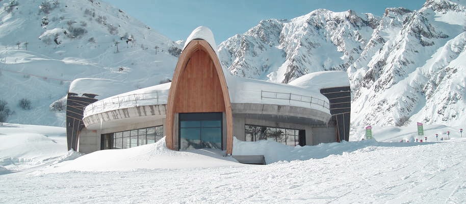 Bâtiment moderne en bois et béton, partiellement enneigé, avec une entrée arquée, sur une piste de ski face à des montagnes.