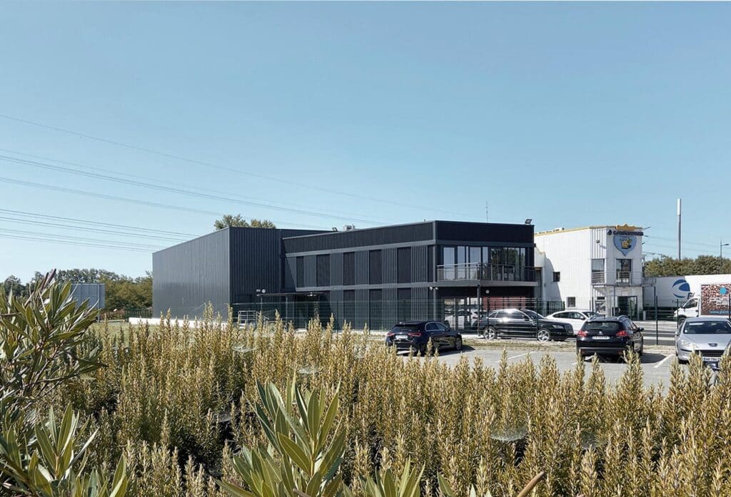 Bâtiments commerciaux: un noir moderne avec balcon, et un blanc LAVATRANS avec camion. Parking ensoleillé et végétation dense.