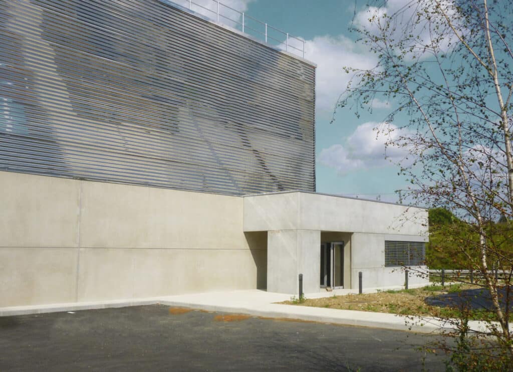 Façade moderne d'un bâtiment en béton et métal avec lattes horizontales. Un bouleau argenté à droite sous un ciel nuageux.